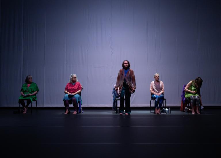 En gästdansare går mot dig. Bakom henne sitter andra gästdansare på stolar på en rad.