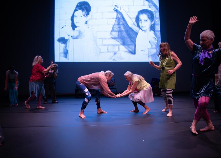 Två gästdansare över 60 år dansar tillsammans med händerna mot varandra omgivna av andra dansare. Bakom dem visas ett äldre  foto av två unga flickor som dansar. 