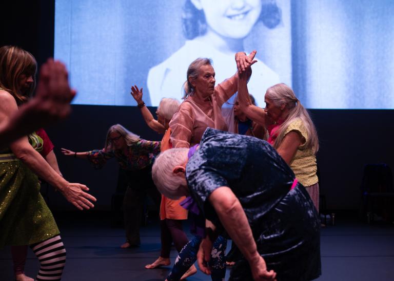 Gästdansare över 60 år dansar på rad på egna vis. Bakom dem visas ett svartvitt porträtt av en ung flicka på en filmduk.