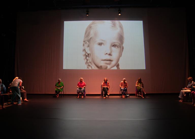 Gästdansare sitter på stolar längs sidorna av en stor scenyta. På en filmduk på bakre väggen projiceras ett svartvitt porträtt på en ung flicka med flätor.