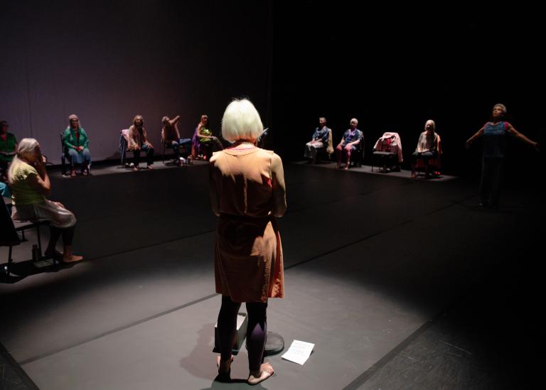 En gästdansare i ljusorange klänning står i ett runt strålkastarljus med ryggen vänd mot dig. Framför henne i en stor fyrkant längs sidorna av en scen sitter de andra dansarna i föreställningen.