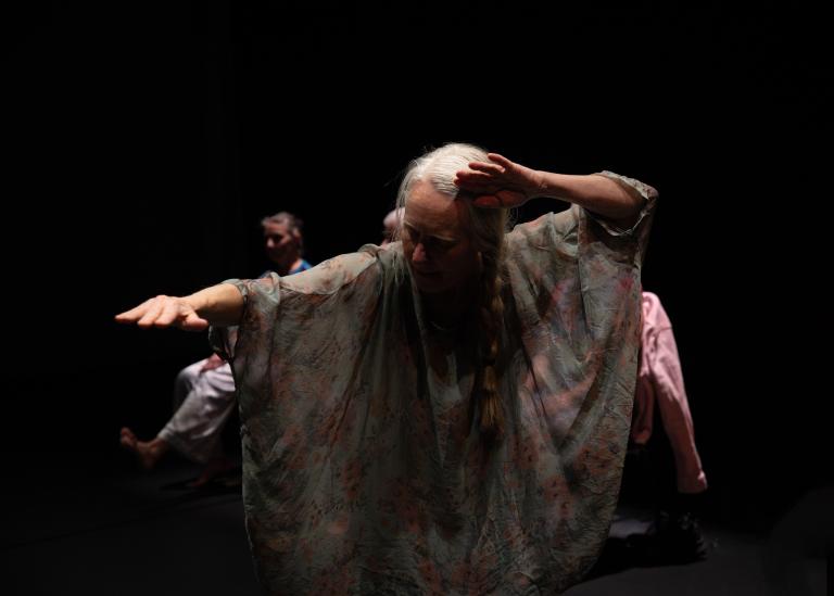 En gästdansare över 60 år klädd i en ljus böljande tunika med håret i en lång fläta sträcker fram ena armen framför sig och lyfter den andra handen bakåt mot sitt öra i dansföreställningen Jag minns mig själv.