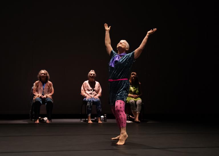 En dansare i lilablå tunika och rosa leggings, med kortklippt grått hår sträcker armarna över huvudet och vänder ansiktet uppåt.