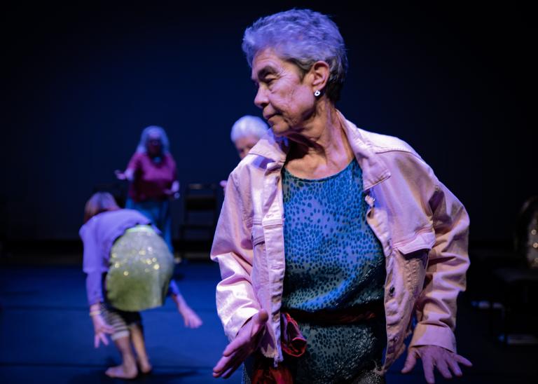 En gästdansare över 60 år med kortklippt hår, klädd i ljusrosa jeansjacka och blå blus dansar framför andra dansare i dansföreställningen Jag minns mig själv.