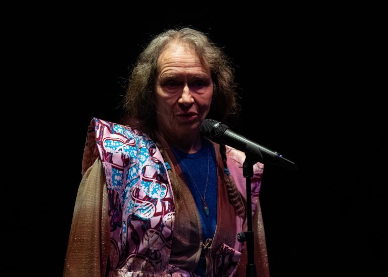 En gästdansare över 60 år med långt lockigt hår och draperade kläder talar i en mikrofon i dansföreställningen Jag minns mig själv.