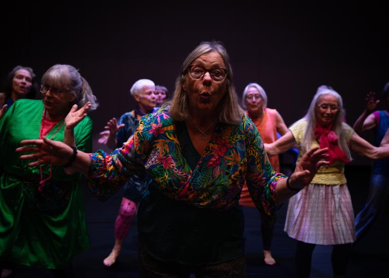 En gästdansare med axellångt hår, klädd i en grön och blommönstrad blus sträcker engagerat ut händerna och formar sin mun som ett O i dansföreställningen Jag minns mig själv.