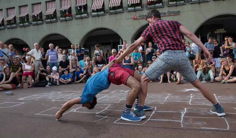 En dansare står bredbent och håller en annan dansare i handen. Den andra dansaren böjer sig bakåt mot marken så att hans huvud och fingrar nästan rör vid marken. 