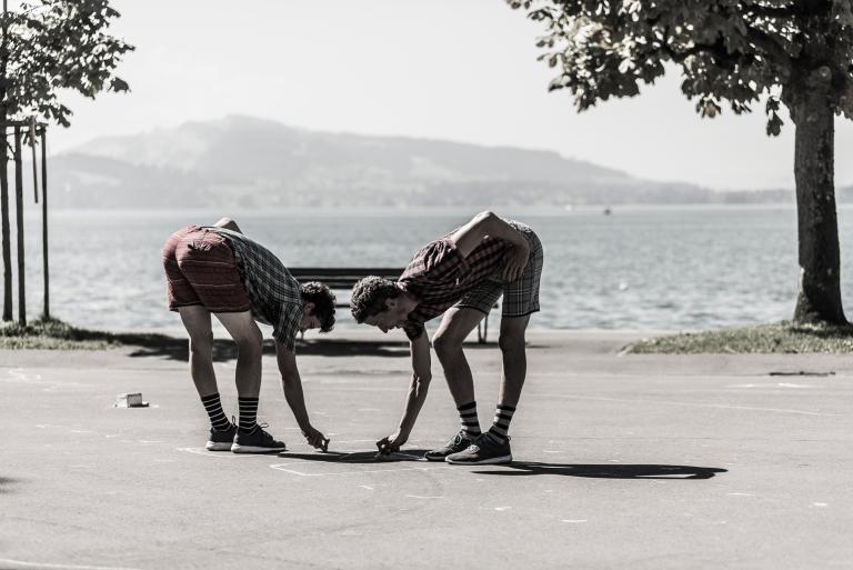 Två dansare i shorts och rutiga skjortor ritar upp en lekhage på marken vid en stor sjö.
