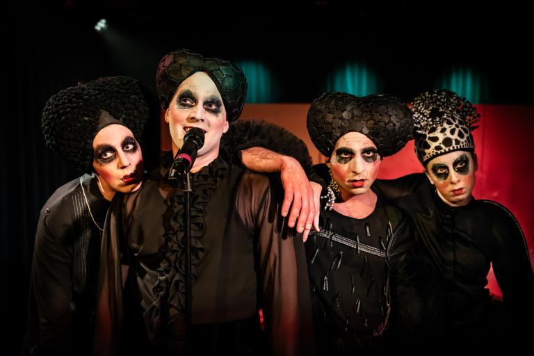 Fyra dansare klädda i svarta kläder och ovanliga hattar strå tillsammans vid en mikrofon. 