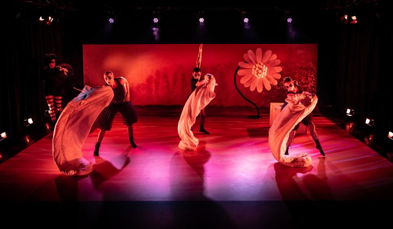 Tre dansare i svarta kläder dansar med tre ljusa skynken på en röd scen med en enorm prästkrage i bakgrunden.