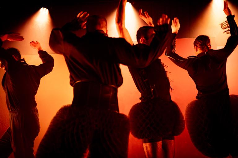 Fyra dansare dansar tillsammans med armarna böjda upp i luften i ett gulrött ljus.