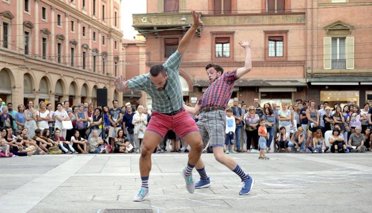 Två dansare i shorts och rutiga skjortor dansar energiskt på ett torg omgivna av publik.
