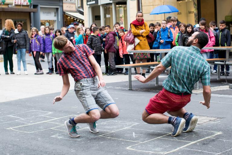Två dansare i shorts och rutiga skjortor balanserar på tå mittemot varandra på en lekhage på en gata.
