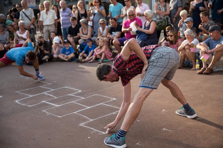 Två dansare i shorts och rutiga skjortor ritar upp en lekhage på marken omgivna av publik.