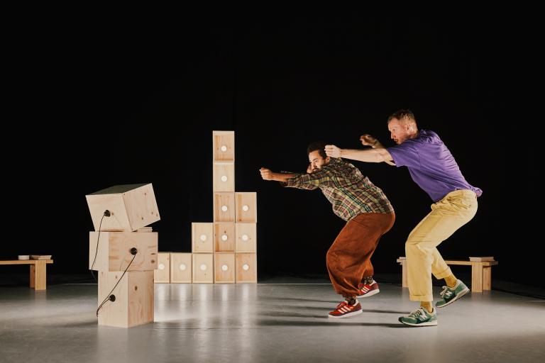 Dansarna Yared och Viktor dansar bredvid varandra bland omålade trälådor med lampor och sladdar i. De siktar tillsammans som om de hade pilbågar mot något vi inte ser.