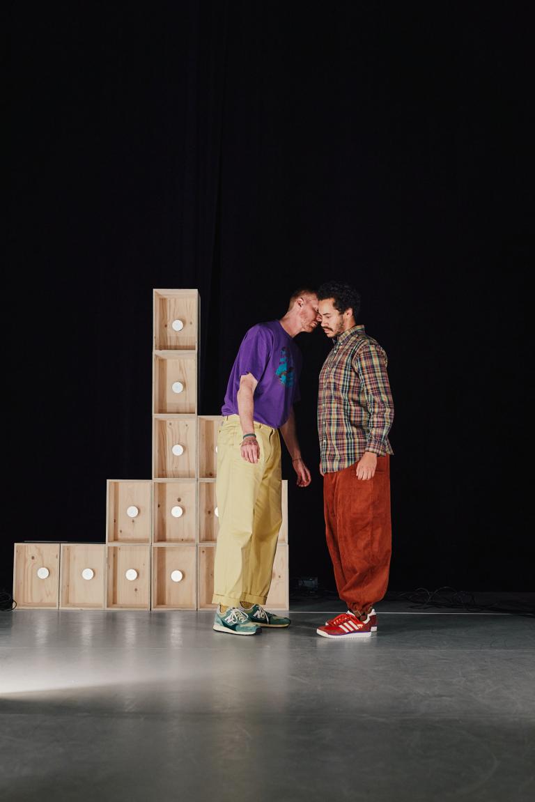 Dansarna Vitkor och Yared står tätt ihop vid staplade omålade träboxar.