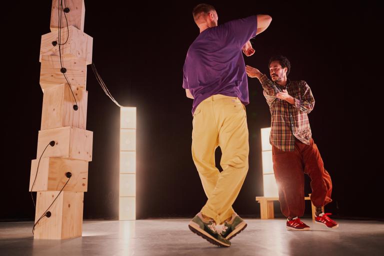 Dansarna Vitkor och Yared dansar med armar och ben korsade bland staplade omålade träboxar som lyser på dem.