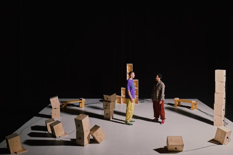 En bild ovaninfrån på en vit ascen där dansarna Viktor och Yared står bland många omålade trälådor som står staplade och ligger huller om buller. 