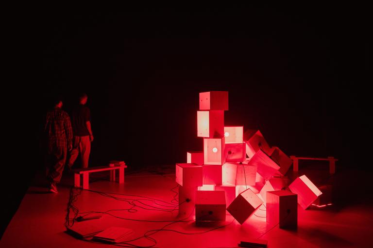 En stor hög av omålade trälådor står staplade på en scen i rött ljus.