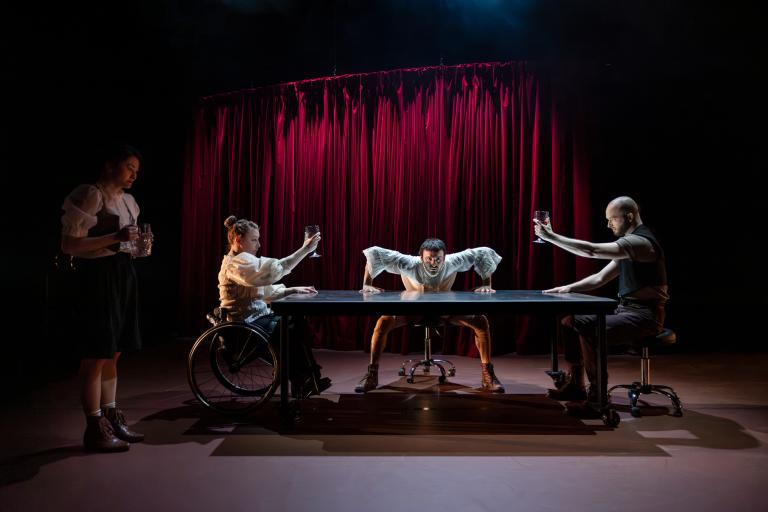 Dansarna MAdeleine och Tomáš sitter mittemot varandra vid ett långt bord. Mellan dom lutar sig dansaren Facundo på bordet och stirrar på dig. I skuggan vid sidan av bordet står dansaren Hazuki med en kanna. Madeleine och Tomáš höjer sina glas som i en skål.