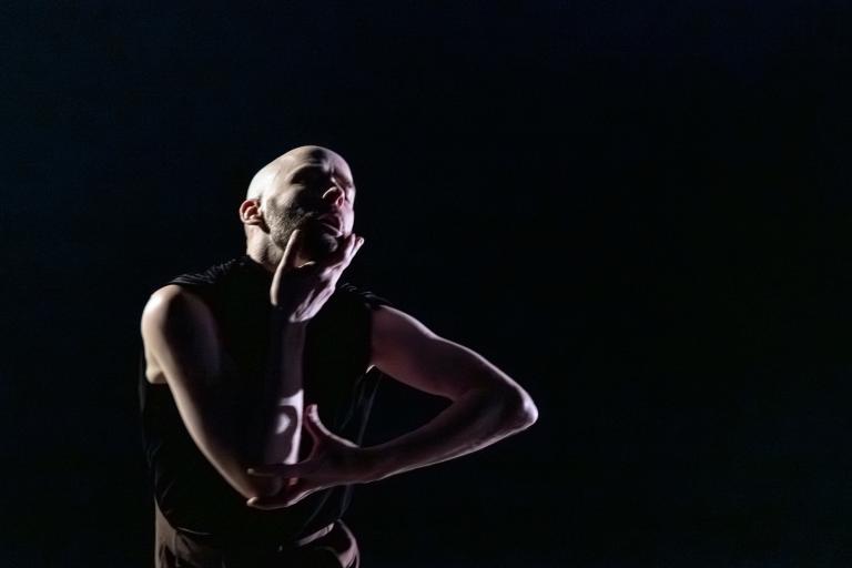 Dansaren Samuel dansar i mörkret belyst från sidan. Han har en svart väst på sig och dansar med armarna böjda i vinklar och en hand på sin haka. 