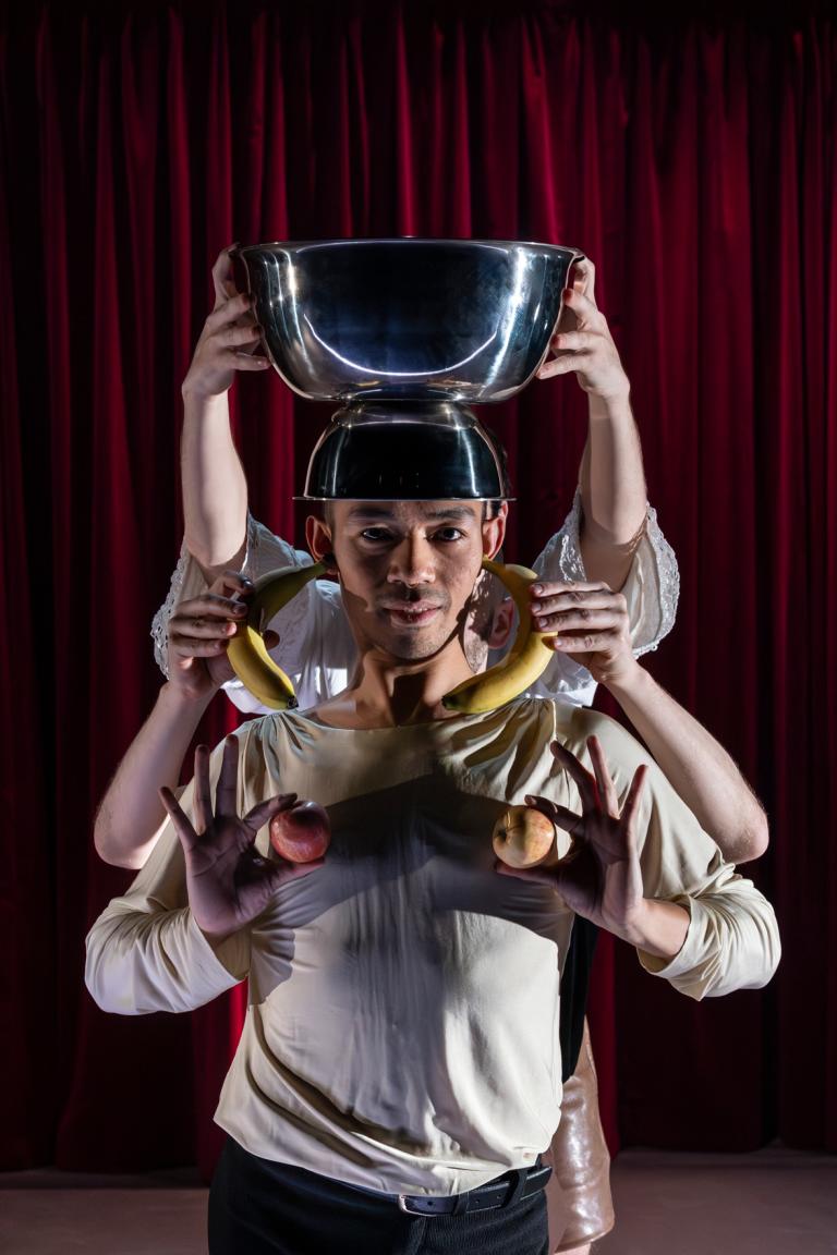 Dansaren Michael håller två äpplen som ett par bröst. Bakom honom håller någon bananer vid hans öron som örhängen och en stor silvrig skål på fot på hans huvud som en hatt. 