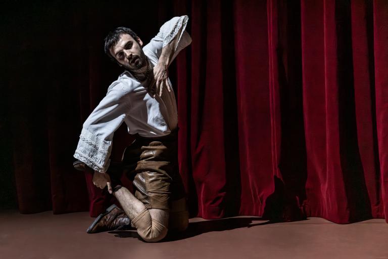 Dansaren Facundo dansar på knä klädd i ljusbruna lädershorts och en broderad skjorta med volanger. Han böjer upp ena foten i luften, vinklar ena armen upp i vinkel och vrider sig mot dig. 