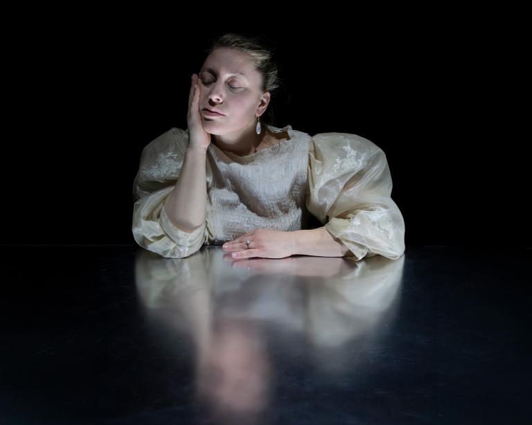 Dansaren Madeleine i en puffig cremefärgad brokadblus vid ett blankt bord. Hon blundar lugnt med hakan i handen.