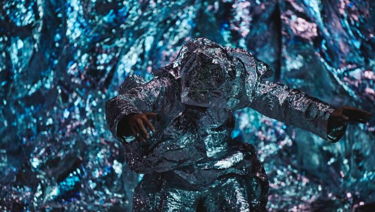 En dansare rör sig framför en glänsande metalisk grottliknande vägg. 