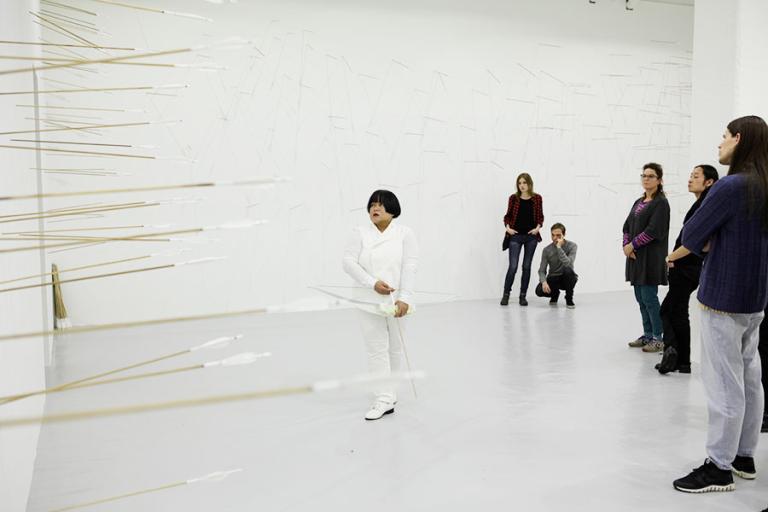 Konstnären Melati Suryodarmo klädd i vitt med en pilbåge står vid en vägg täckt av pilar. Bakom henne står publik.