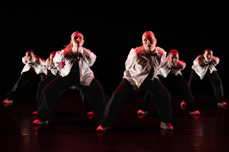 Sex unga dansare i svrat byxor och vita skjortor dansar i en triangelform och lutar sig fram mot dig.