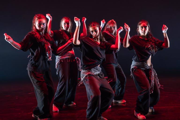 Fem dansare klädda i svart dansar tillsammans mot dig med armarna böjda och händerna knutna.