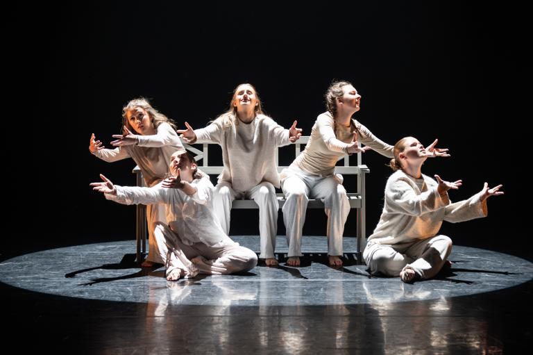 Fem dansare klädda i vitt sitter med utsträckta armar som i en vädjan på och runt en bänk.