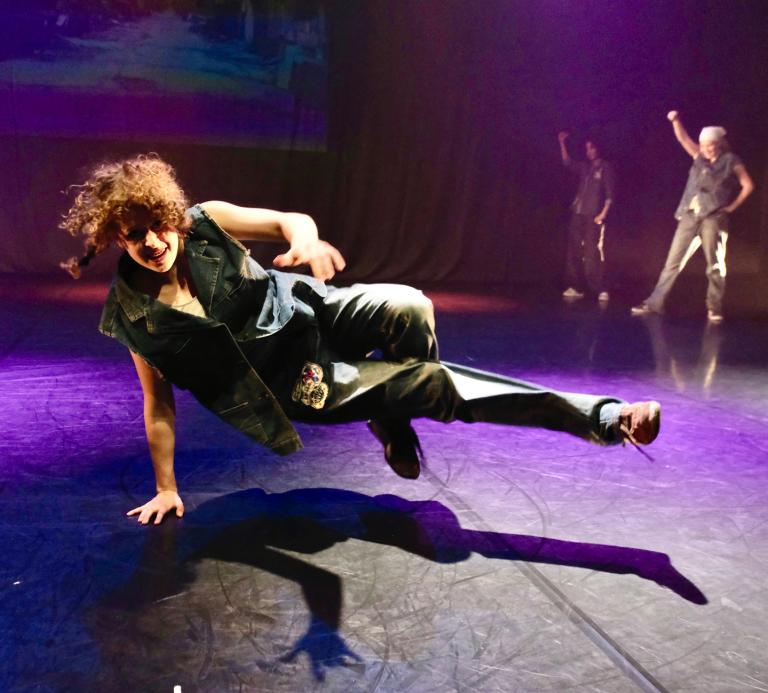 En dansare med långt brunt hår kläd i jeans gör en dansrotation på golvet balanserande på en hand.