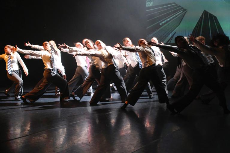 En stor grupp dansare i vita skjortor och svarta byxor dansar på rad och sträcker ut ena armen synkroniserat.