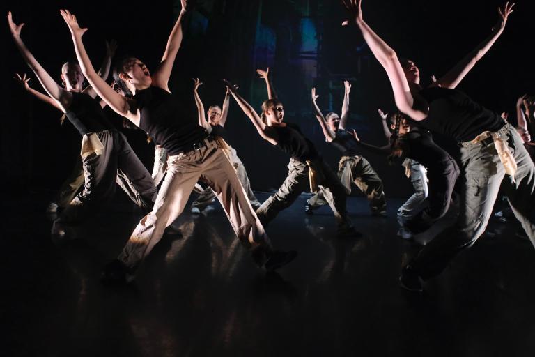 En stor grupp dansare i ljusa byxor och mörka linnen dansar bredbent med armarna sträckta ovanför sig.