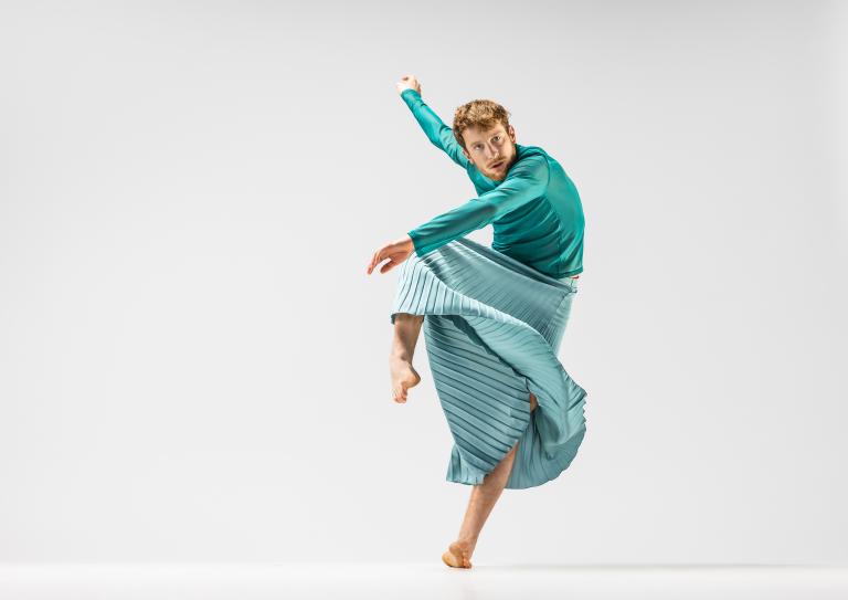 Skånes Dansteaters dansare Jure Gostinčar klädd i turkost dansar på ett ben med andra knät och armarna böjda vid sidan.