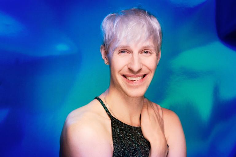 Artisten Oscar Pierrou Lindén med kort vitt hår och glittrigt svart linne framför en blåskimrande bakgrund.