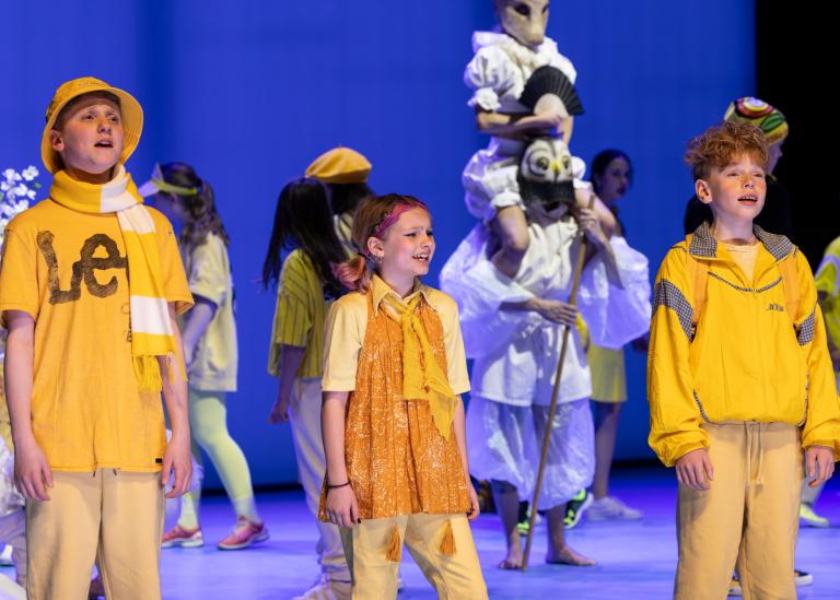 Tre unga barnsångare klädda i moderna gula kläder sjunger. Bakom dem rör sig andra barn i gult och en dansare som bär en annan dansare på sina axlar i Skånes Dansteaters dansföreställning Out of the Blue.