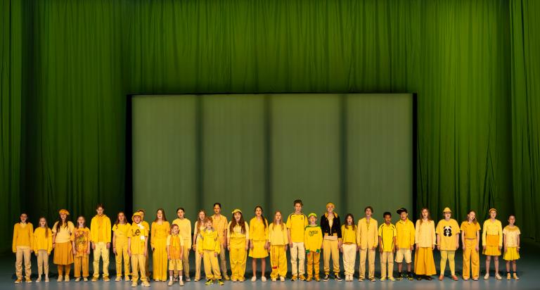 Malmö OPeras barnkör klädda i olika gula kläder står längs scenkanten och sjunger i ett grönt rum i Skånes Dansteaters dansföreställning Out of the Blue. 