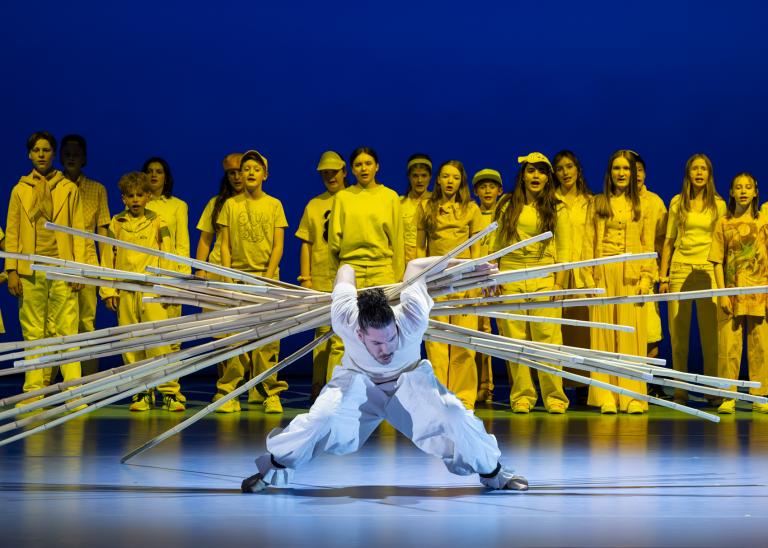 Dansaren FAbrizio di Franco böjer sig fram under ett stort antal långa träpinnar som han håller på ryggen med sina armar. Bakom honom sjunger Malmö Operas barnkör klädda i gula kläder i Skånes Dansteaters dansföreställning Out of the Blue.