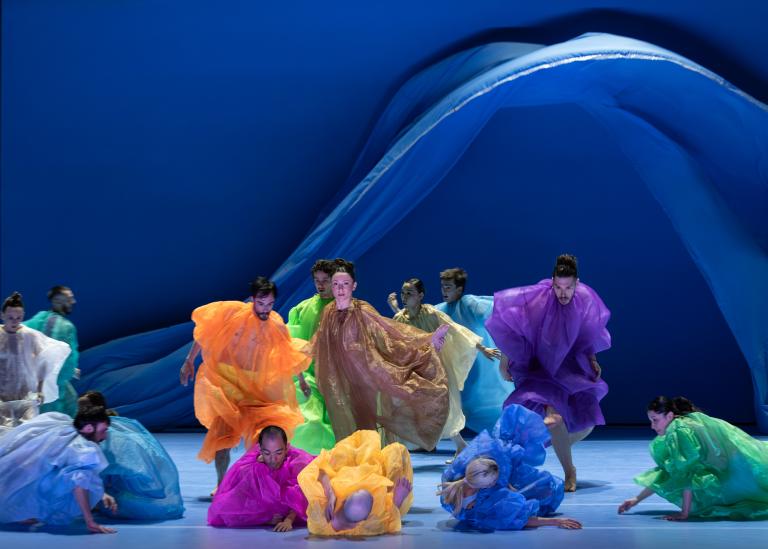 Dansare i skira overaller i klara färger dansar medan stora blå skynken faller bakom dem i Skånes Dansteaters dansföreställning Out of the Blue.
