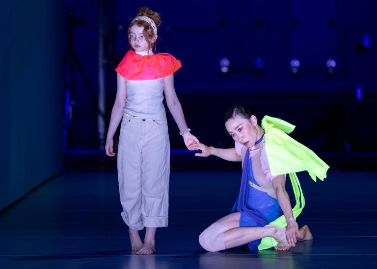 En ung flicka med rött uppsatt hår och ljusa kläder håller dansaren Jing Yi Wang i handen i Skånes Dansteaters dansföreställning Out of the Blue.