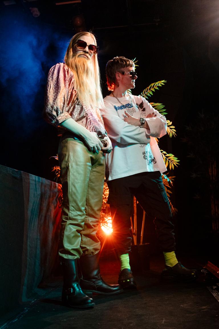 Två dragkings med solglasögon står på en scen i strålkastarljuset.