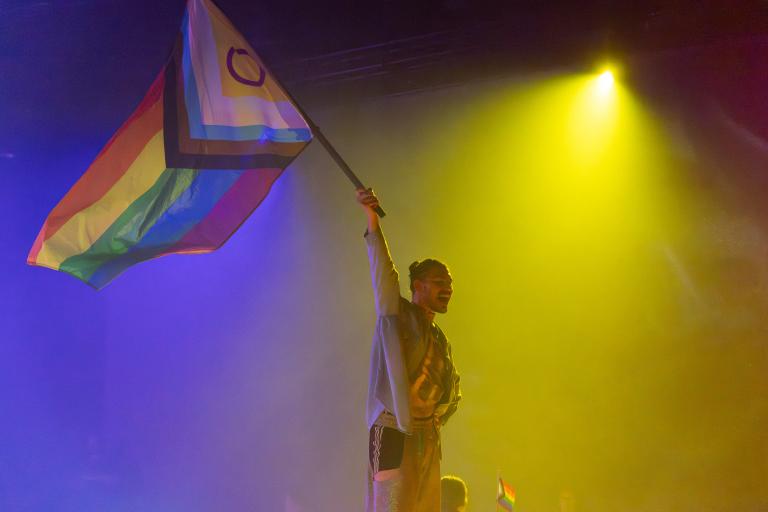 Dansaren Fabrizio klädd i silvriga chaps sveper en Pride-flagga genom luften i Skånes Dansteaters dansföreställning Indigo Flamingo.