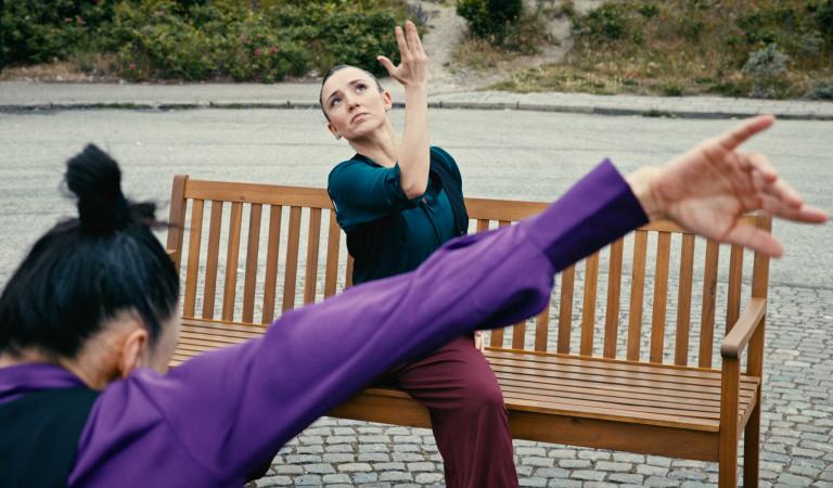 En dansare sitter på en träbänk och sträcker ena handen uppåt i en uttrycksfull rörelse. I förgrunden syns en annan dansares utsträckta arm. Föreställningen utspelar sig utomhus på en stenlagd plats.