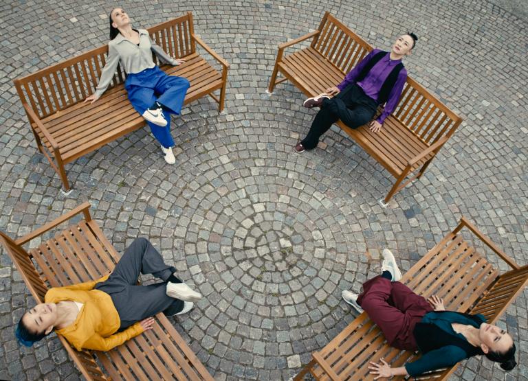 Fyra dansare sitter på varsin träbänk placerade i en cirkel på en kullerstensbelagd yta. Fotot är taget ovanifrån och skapar ett symmetriskt mönster.