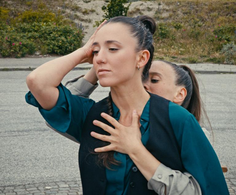 Två dansare står tätt intill varandra i en koreograferad omfamning. Den ena vilar handen mot sitt huvud medan den andra omsluter henne bakifrån.