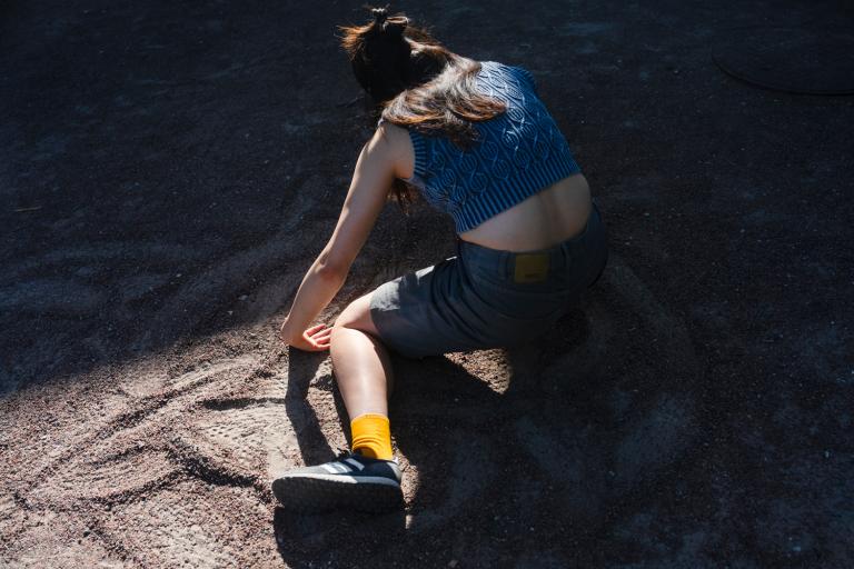 Dansaren Amanda sitter på marken med benen på sidan. Hon är klädd i en blå kort topp utan ärm, shorts, gula strumpor och sneakers. Hondrar sin hand i gruset på marken och följer mönster som hon gjort.