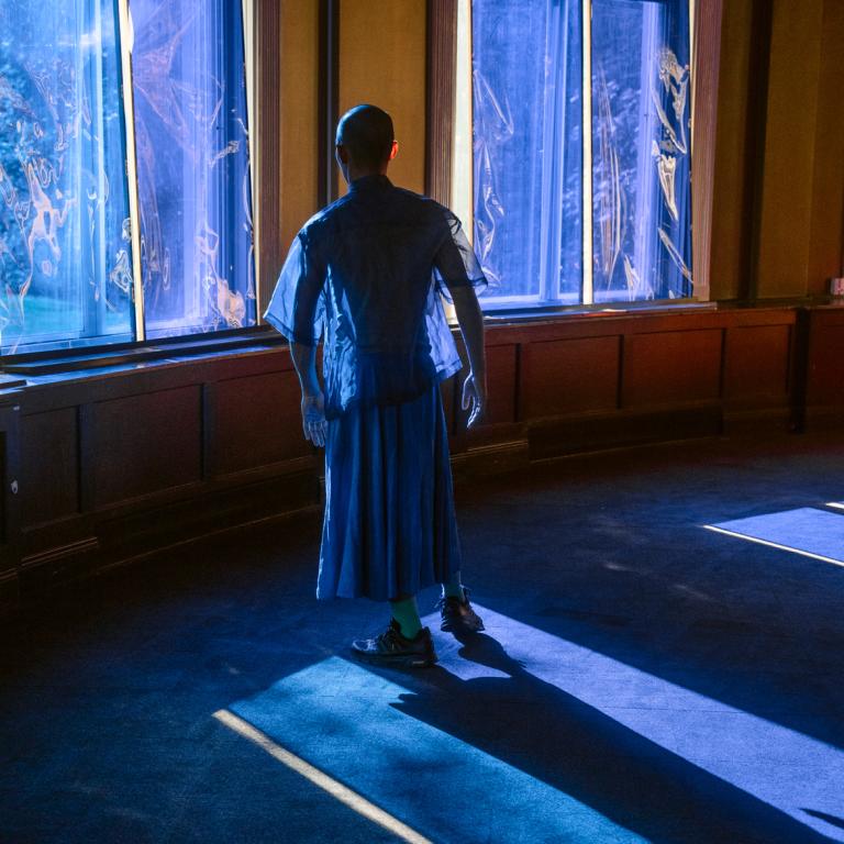 Dansaren Michael står klädd i skira blå byxor och skjorta intill ett fönster täckt med blå platsfilm i Skånes Dansteaters föreställning The Upheaval.